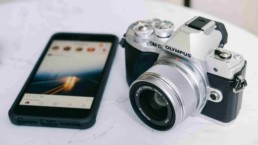 Telefon z włączonym ekranem i Instagramem oraz aparat marki Olympus na marmurowym stole